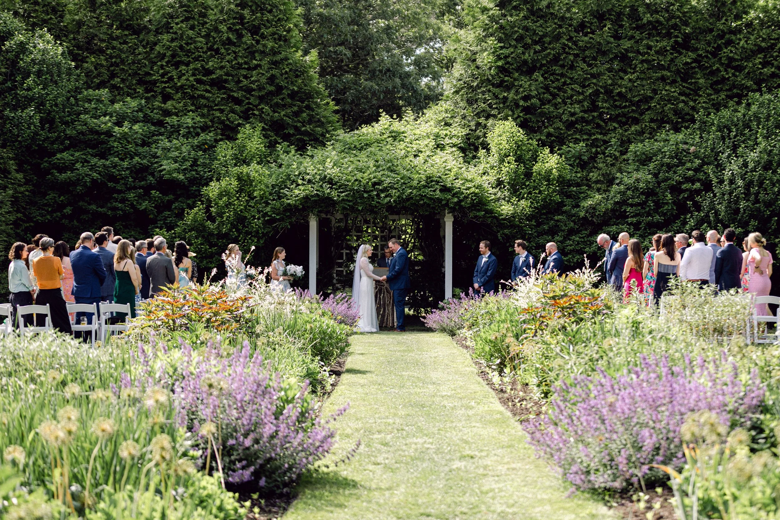 A wedding ceremony outdoors in the English garden at Lambert's Cove Inn on Martha's Vineyard with a crowd of guests, a couple exchanging vows under a decorated canopy, surrounded by blooming lavender and green trees.