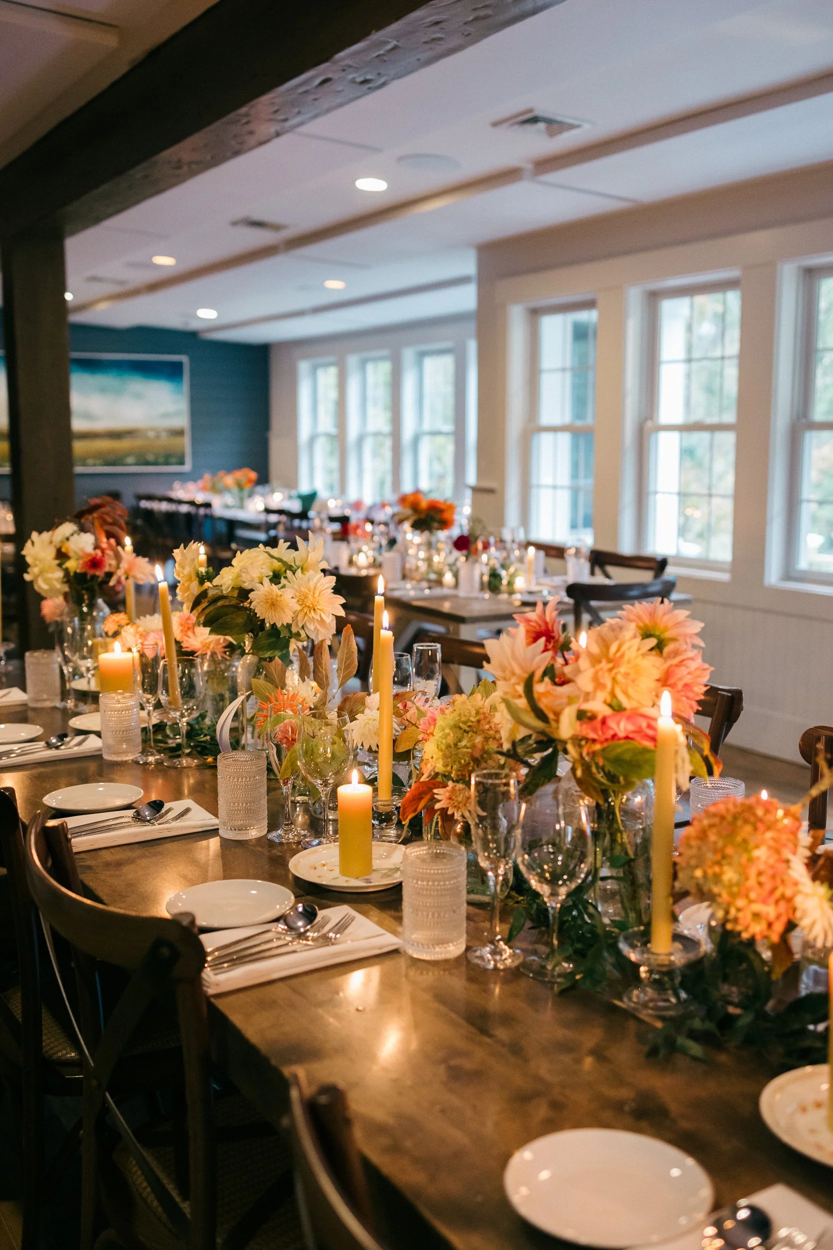 A dining table at Lambert's Cove Inn on Martha's Vineyard decorated with bouquets of pink and white flowers and lit candles in glass holders, set for a meal with plates, silverware, and glasses, in a bright room with large windows.