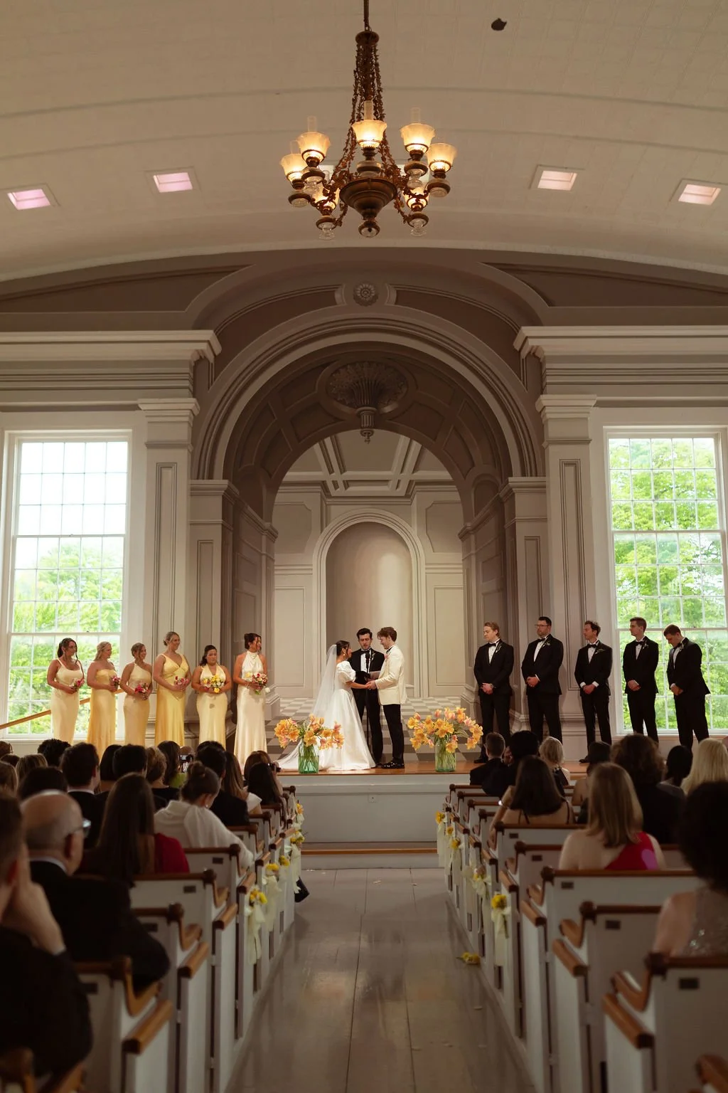 A wedding ceremony taking place in a large, elegant church with tall windows, a chandelier, and ornate architecture. The bride and groom are exchanging vows at the altar, surrounded by bridesmaids in yellow dresses, groomsmen in black tuxedos, and an