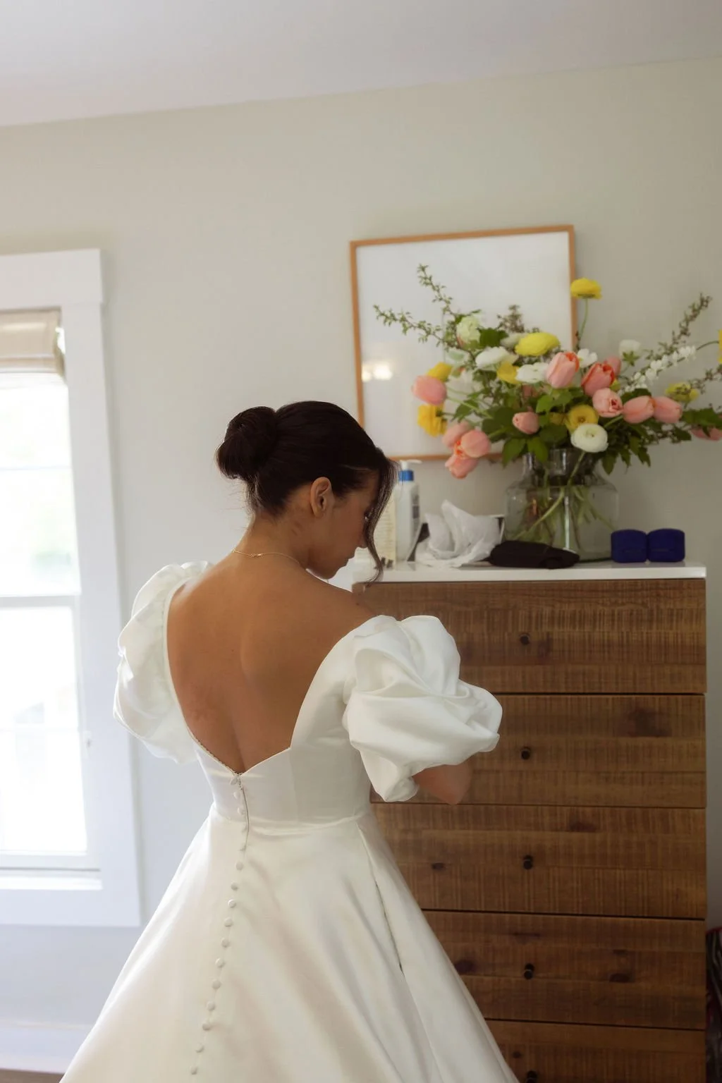 A woman in a white wedding dress with puffed sleeves and buttons down the back, standing in a room with a large vase of pink, white, and yellow flowers on a wooden dresser at Lambert's Cove Inn on Martha's Vineyard.