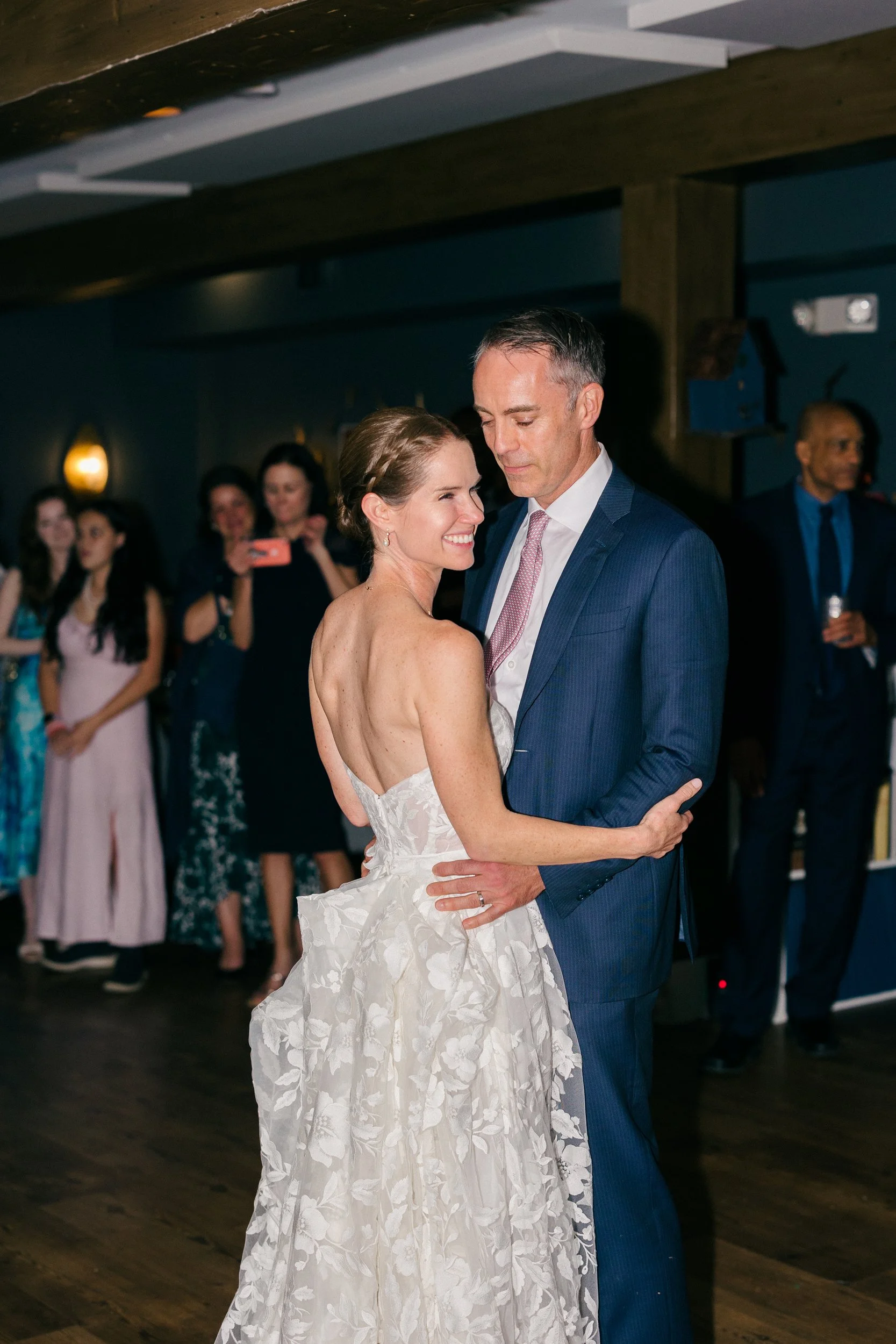 A bride and groom dance closely at their wedding reception at Lambert's Cove Inn on Martha's Vineyard, with guests watching and taking photos in the background.