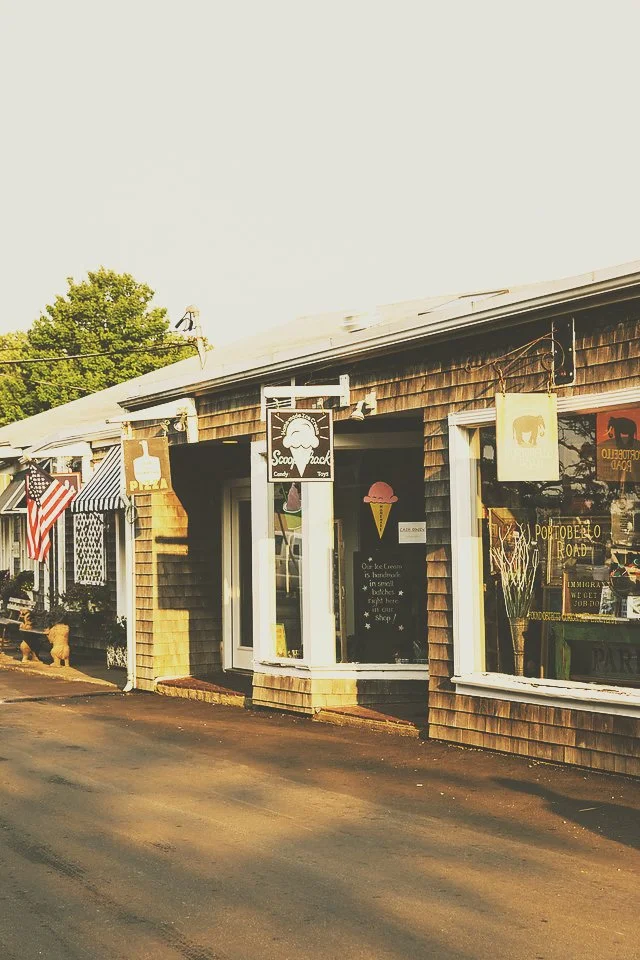 Small storefront shop with wooden exterior, American flag, and signs advertising ice cream, toys, and local shop items. There are front windows showcasing various items inside.