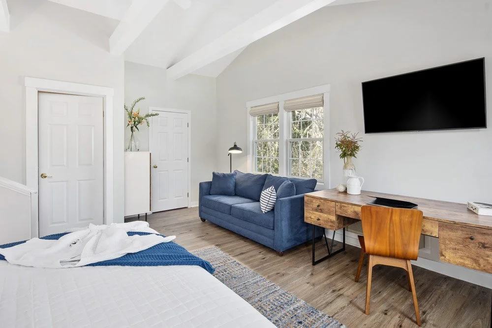 A bedroom with a bed, a blue sofa, a wooden desk with a chair, and a wall-mounted TV, with windows allowing natural light.