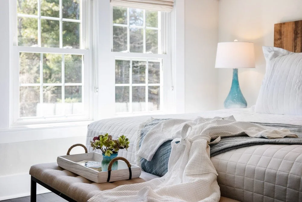 A guest room at Lambert's Cove Inn on Martha's Vineyard with a large window, white comforter, pillows, a blue lamp, and a wooden headboard. There is a bench at the foot of the bed with a tray and a small plant on it.