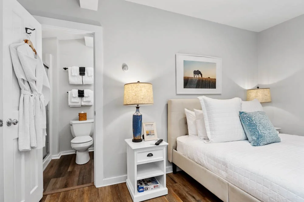 A modern bedroom with a white bed, a nightstand with a lamp, and a framed horse photograph on the wall. An open door reveals a bathroom with towels and a robe.