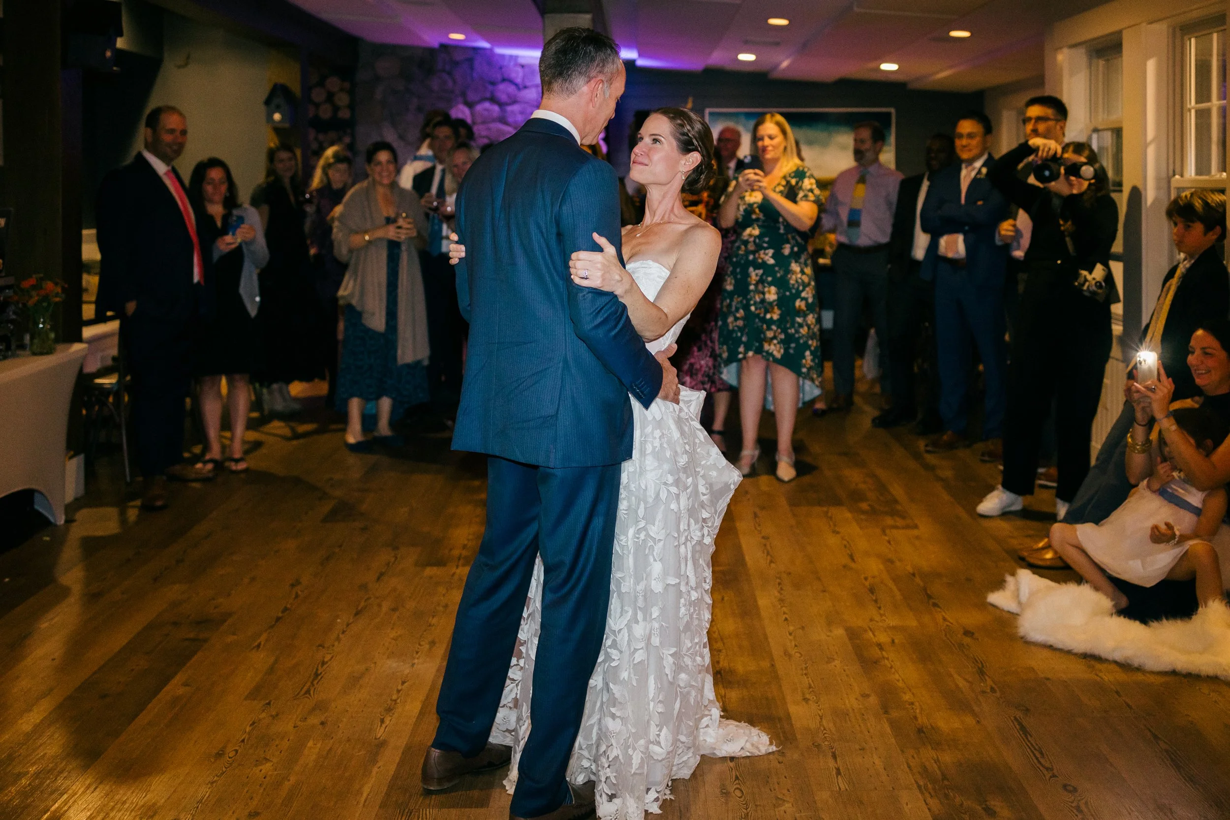 A bride and groom dance together during their wedding reception at Lambert's Cove Inn on Martha's Vineyard, surrounded by seated and standing guests in a warmly lit room.