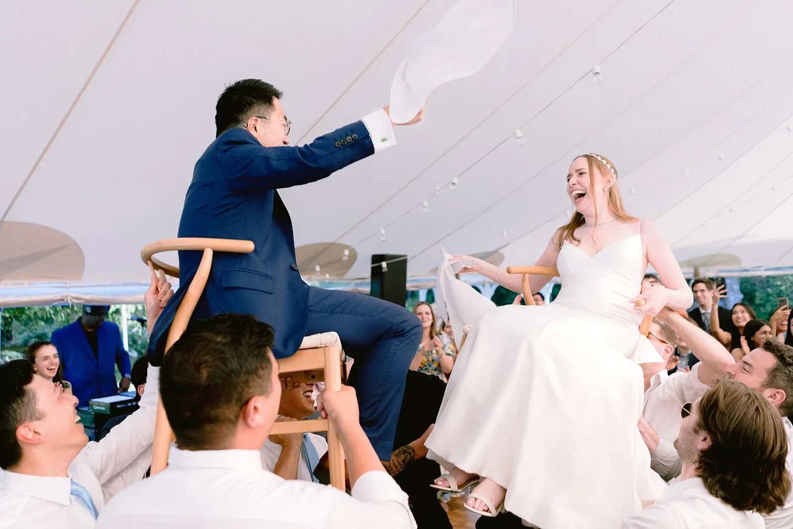 A bride and groom are riding on chairs at their wedding reception at Lambert's Cove Inn. The groom, dressed in a blue suit, is high-fiving the bride, who is wearing a white wedding dress. They are surrounded by smiling guests.