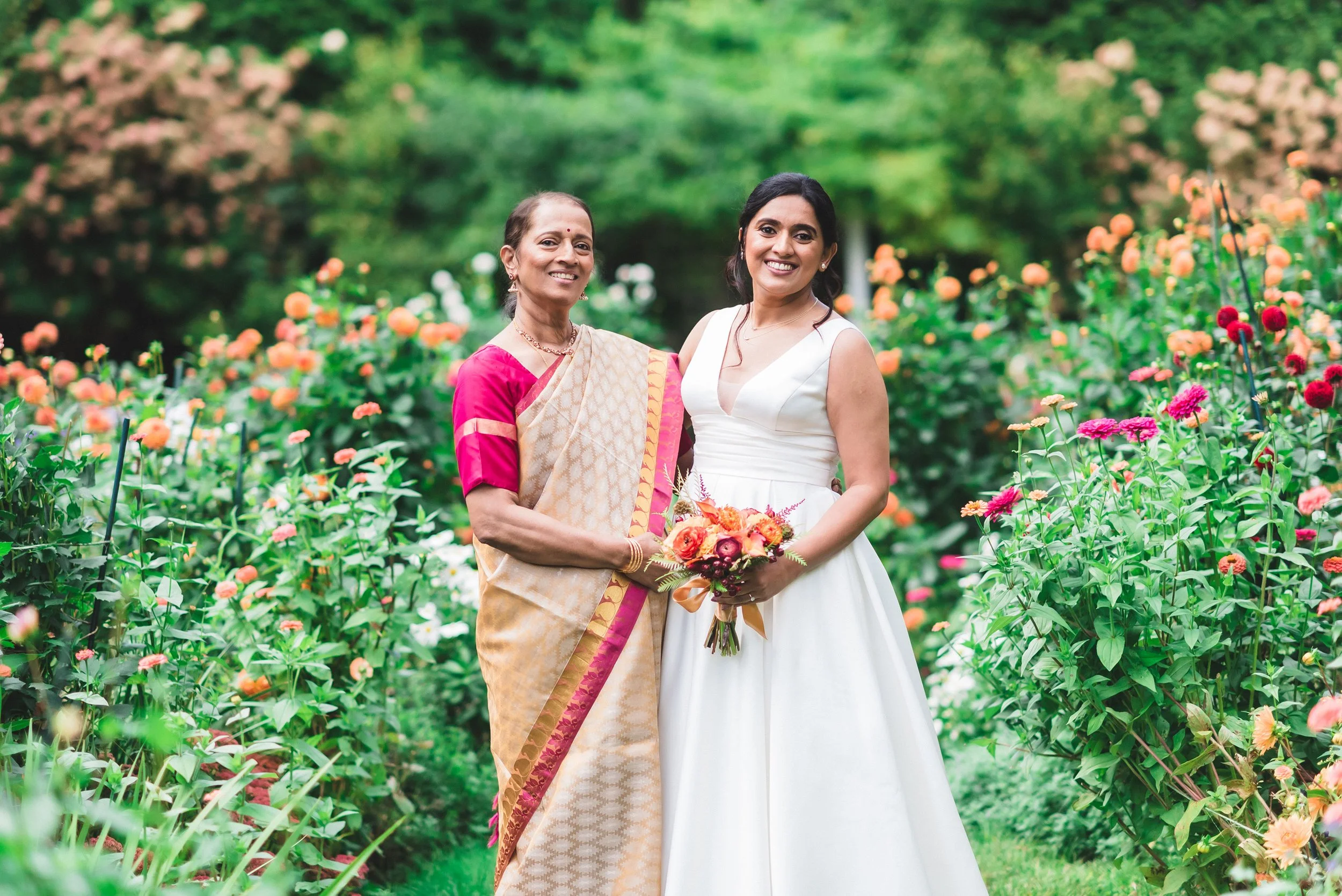 Two women smiling and posing in a garden at Lambert's Cove Inn on Martha's Vineyard with colorful flowers. One woman is wearing a traditional sari and the other a white dress, holding a bouquet of flowers.