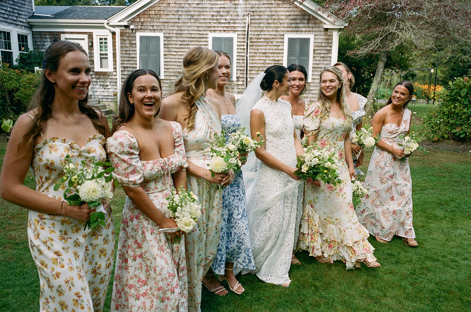 A group of women in floral dresses standing on a lawn, holding bouquets, during a wedding reception at Lambert's Cove Inn on Martha's Vineyard.