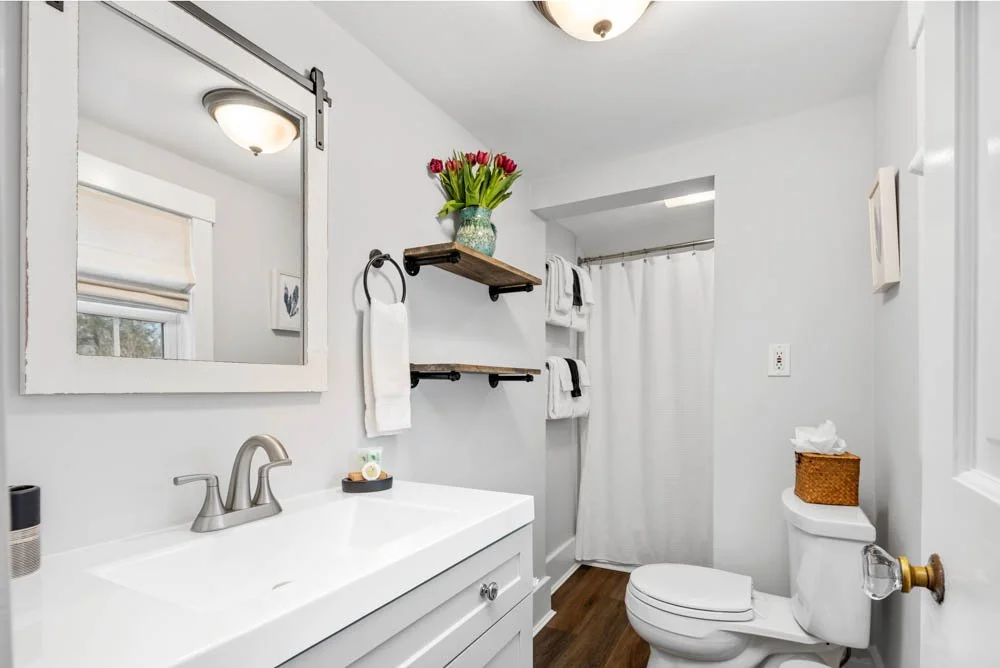 Clean bathroom with white walls, a white vanity with a silver faucet, a wall mirror, a toilet with a tissue box on top, wooden floating shelves with a vase of red tulips and towels, a window with a white shade, and a white shower curtain.