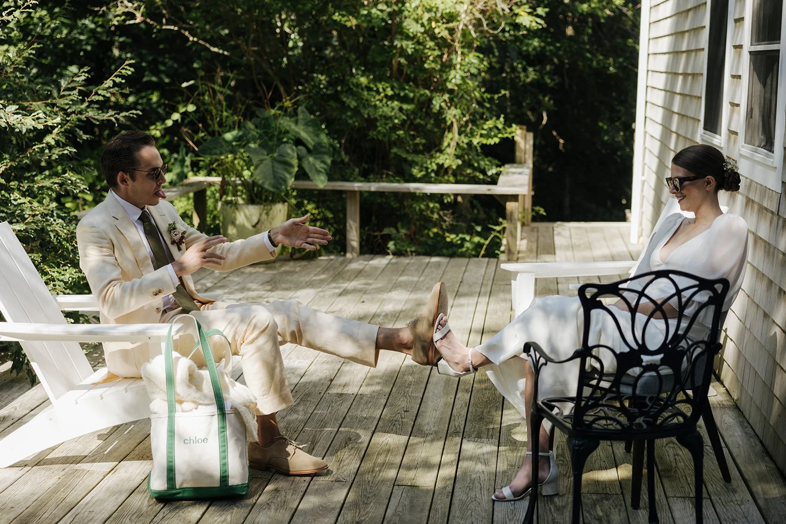 A man and woman in white outfits sitting on a wooden porch. The man is wearing sunglasses and a cream-colored suit, stretching his leg with a shoe, while the woman is smiling, sitting on a white chair, wearing glasses and white heels. They are surrou