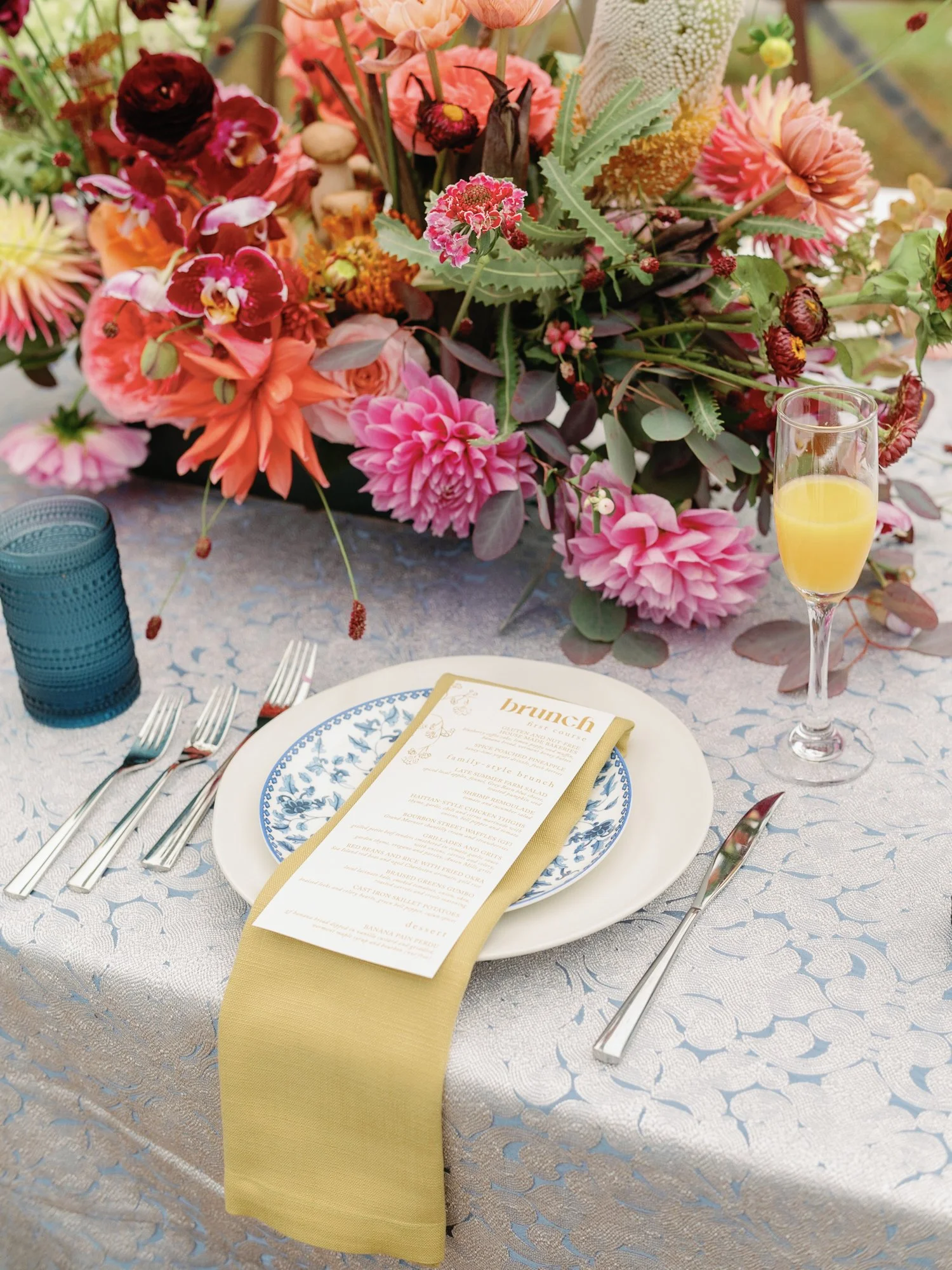 A table setting with a white and blue patterned plate, gold-rimmed napkin, silverware, a menu card, a glass of orange juice, and a large floral centerpiece with pink, red, and peach flowers on a table covered with a light-colored patterned tablecloth