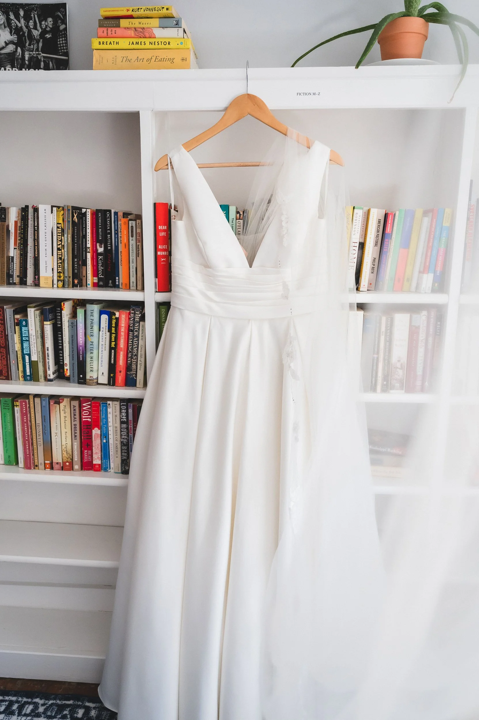 A white dress hanging on a wooden hanger against a white bookshelf filled with books, with a potted plant on top of the shelf, at Lambert's Cove Inn on Martha's Vineyard.