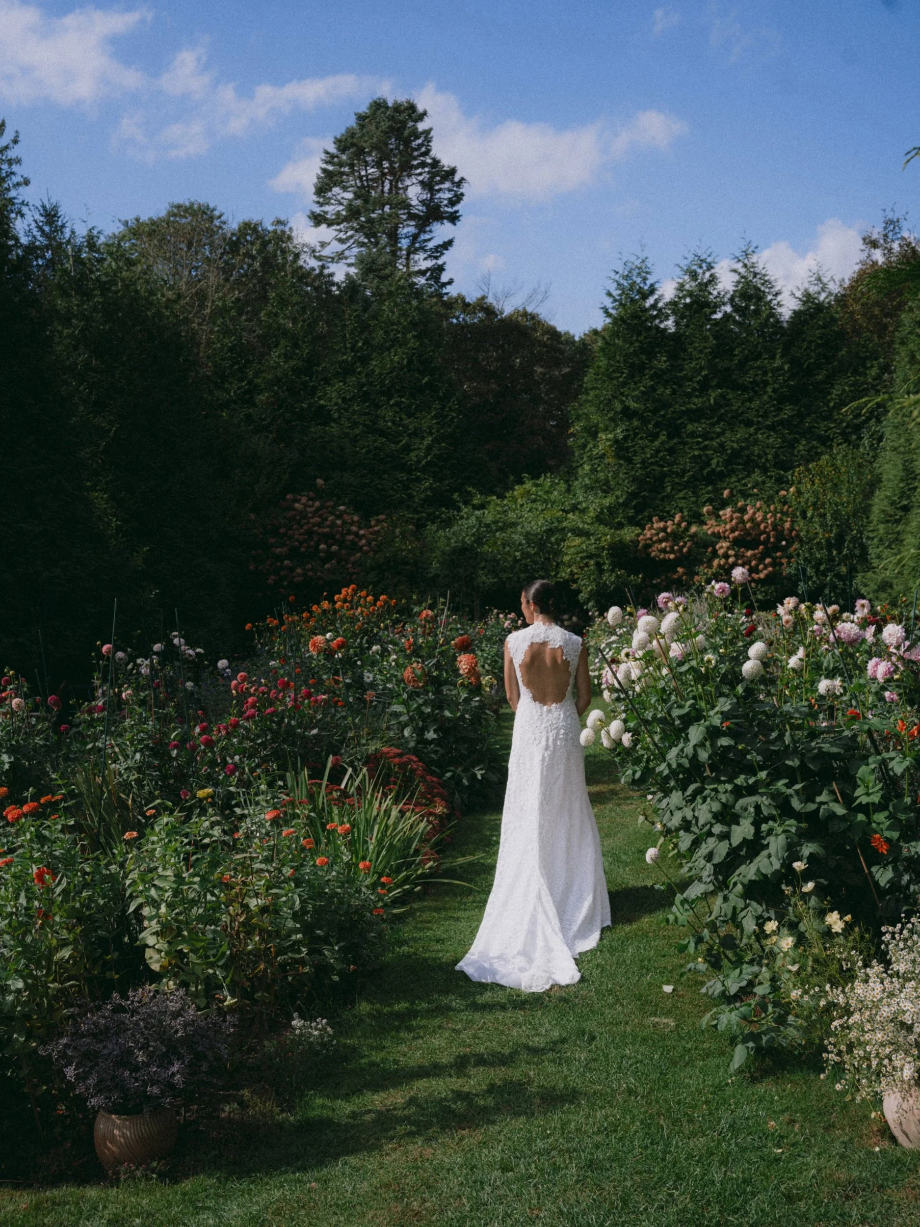 A woman in a long white wedding gown standing in a lush garden surrounded by colorful flowers, with tall trees and a blue sky in the background.