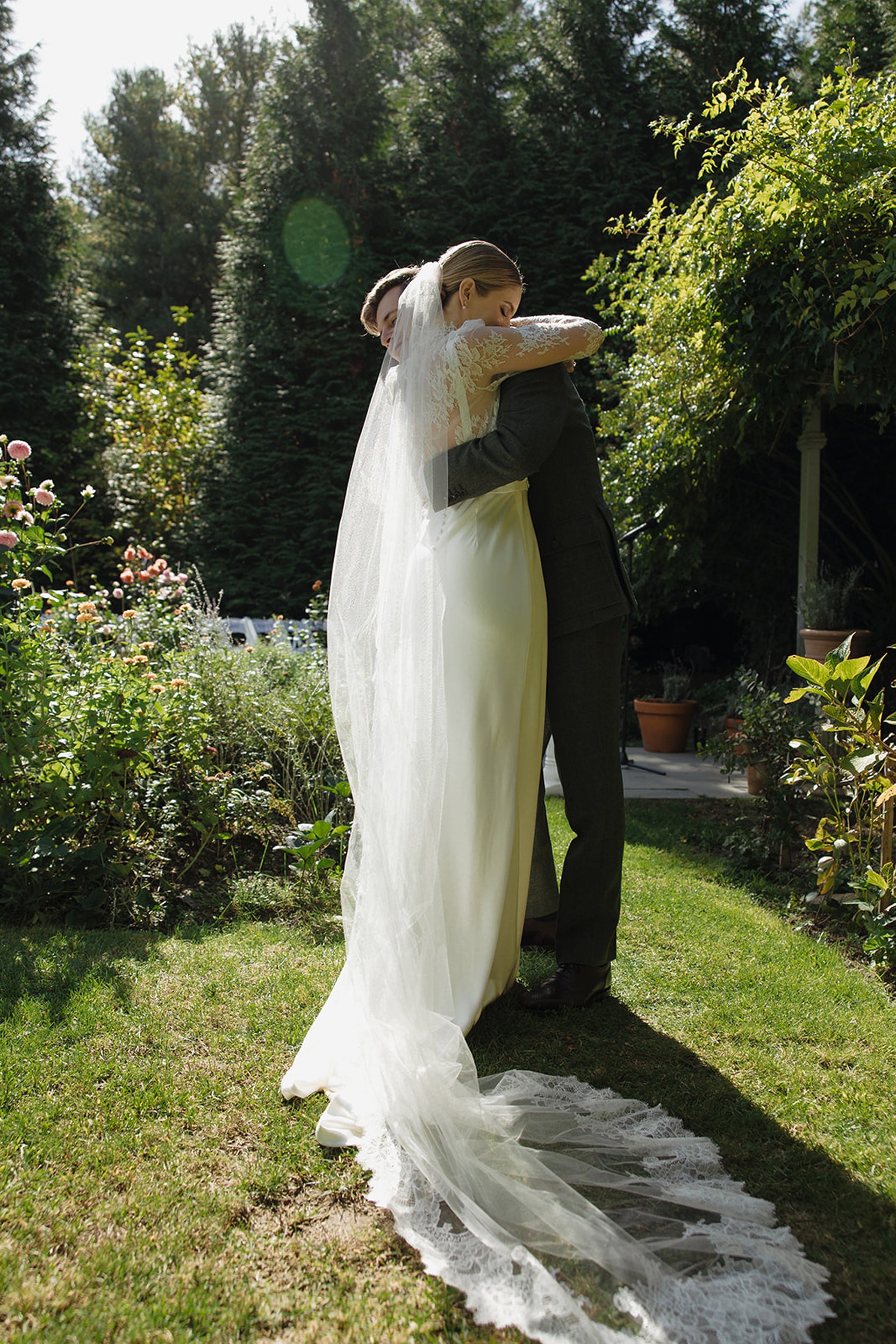 A bride and groom hugging outdoors in a garden, with trees and plants around them on a sunny day.