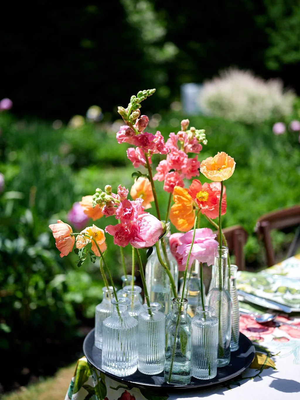 Pink, orange, and yellow poppies arranged in glass bottles on a round tray on a table outdoors before a wedding at Lambert's Cove Inn on Martha's Vineyard.