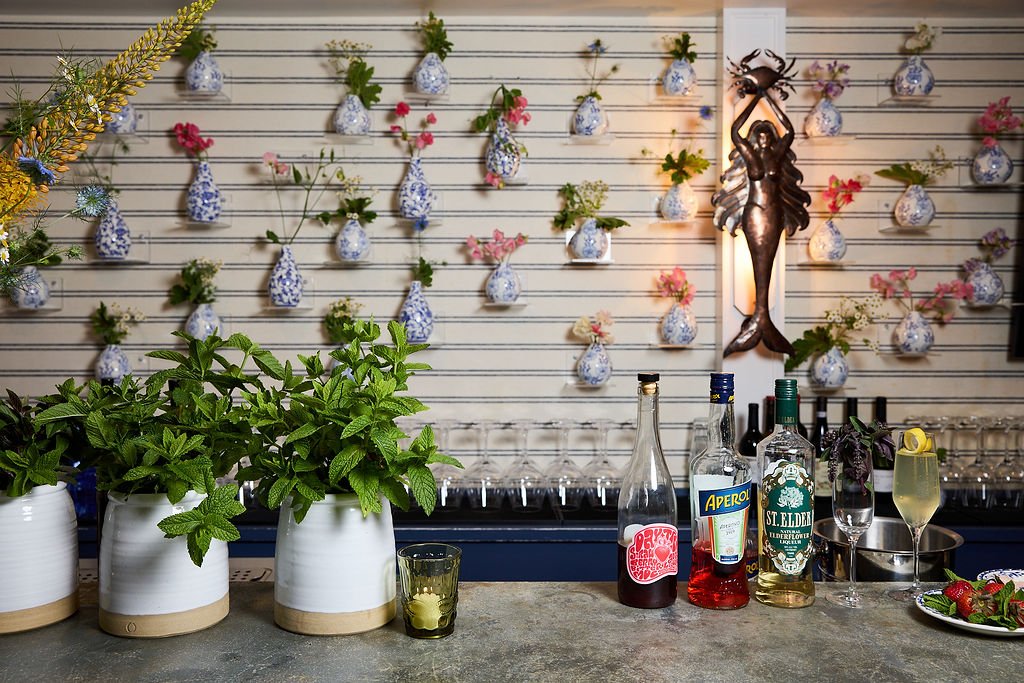 Inside at Mermaid Bar at Lambert's Cove Inn on Martha's Vinyard with potted plants, bottles of liquor, a glass of cocktail, and a tray of strawberries, with a decorative wall featuring hanging vases with flowers and a metal mermaid wall art.