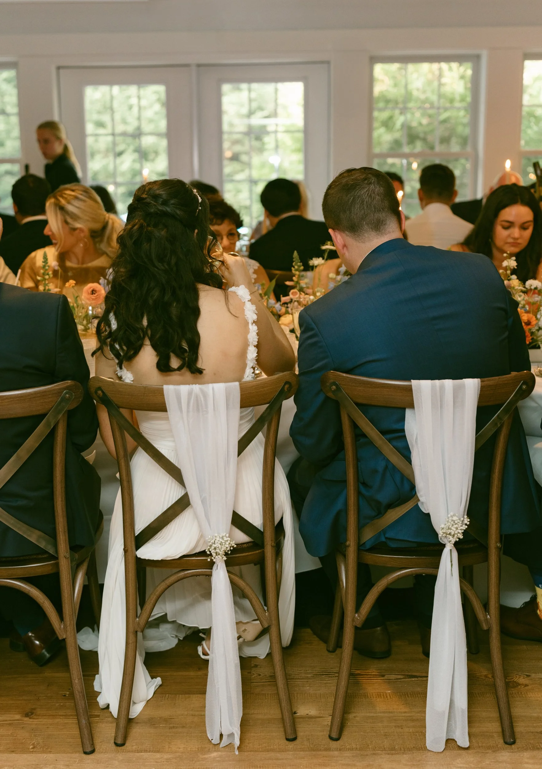 People seated at a wedding reception table at Lambert's Cove Inn on Martha's Vineyard, with a bride and groom in the foreground, decorated with flowers and white drapes on chairs, inside a bright room with large windows and greenery outside.