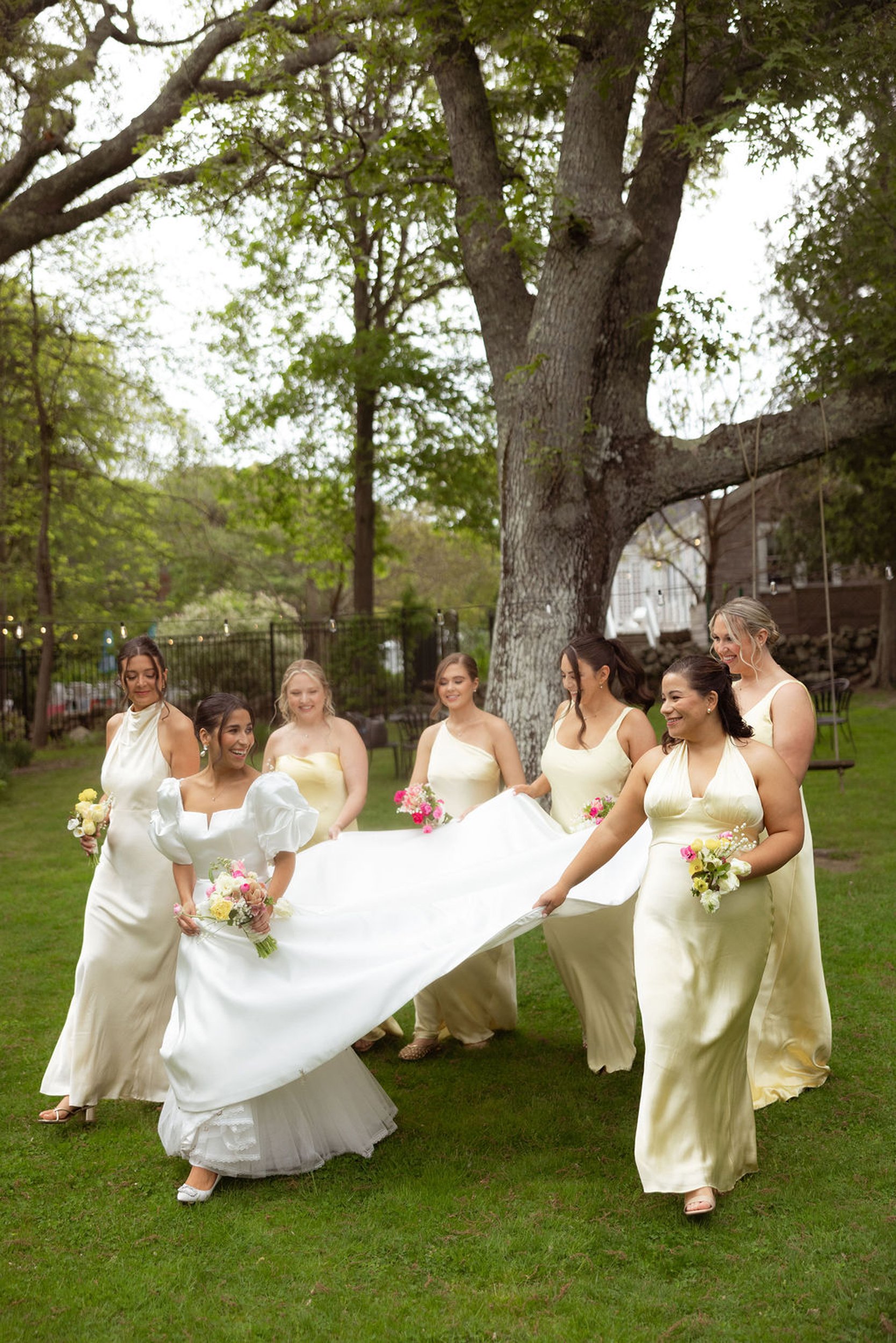 Group of women in light yellow dresses holding bouquets, walking outdoors on grass, at a wedding at Lambert's Cove Inn on Martha's Vineyard, with trees and string lights in the background.