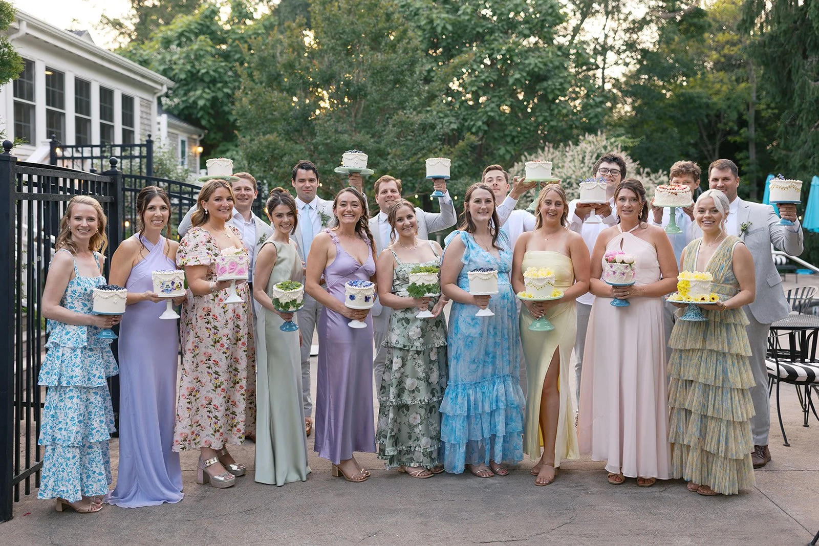 A group of wedding guests standing together outdoors at Lambert's Cove Inn on Martha's Vineyard, each holding a decorated cake. The guests are dressed in pastel-colored dresses and suits, and the background features trees and a house.