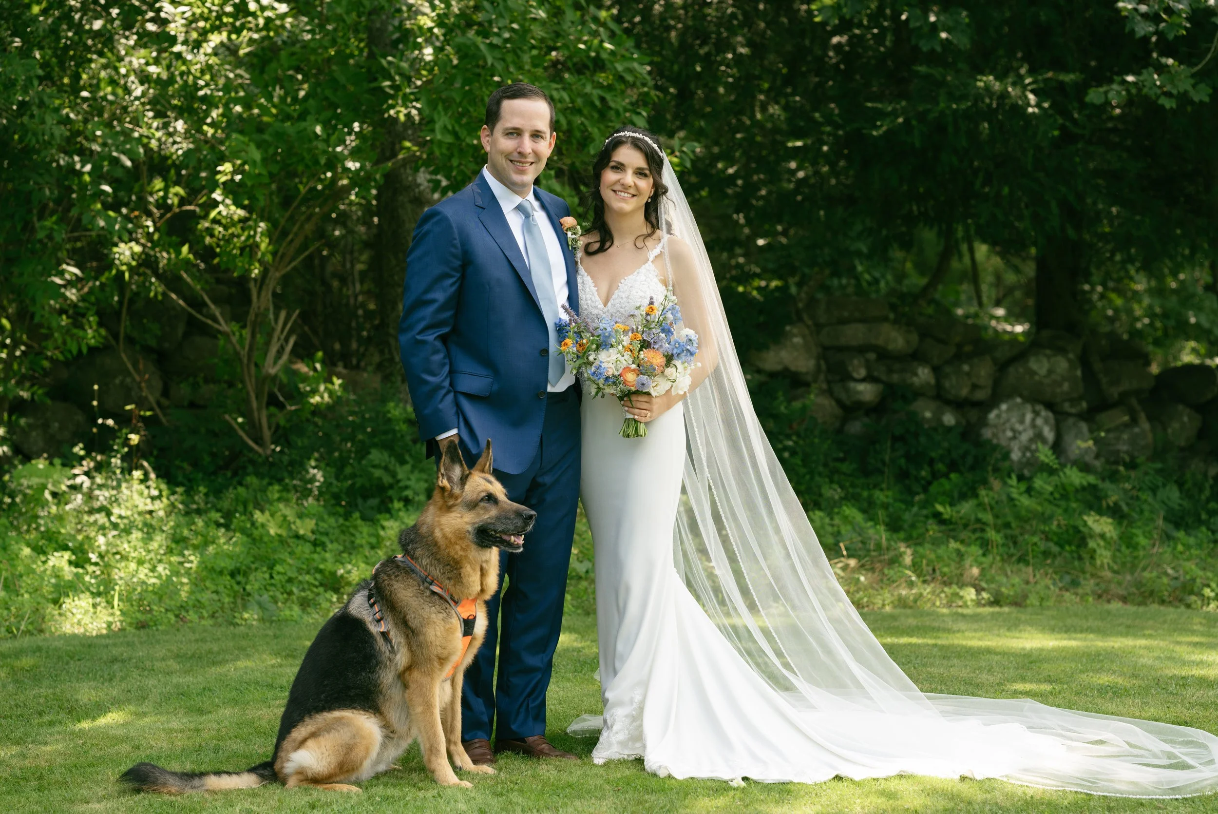 A bride and groom posing for a wedding photo outdoors with a dog sitting in front of them, surrounded by green trees and grass during a wedding at Lambert's Cove Inn on Martha's Vineyard.