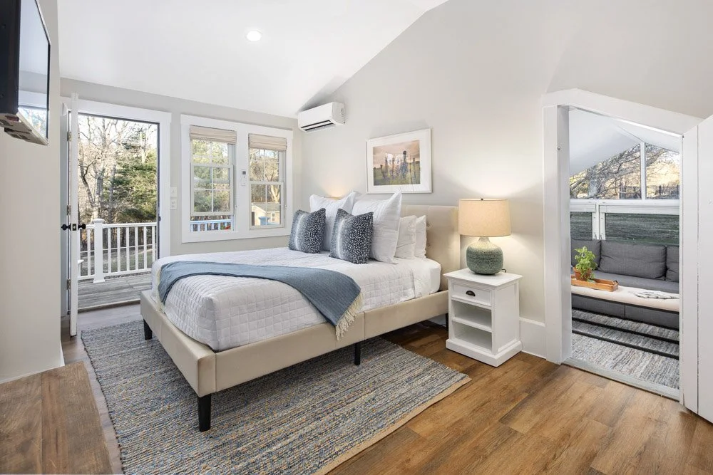 Bright bedroom with a large bed, white bedding, gray pillows, and a blue throw blanket. There's a window with a door leading to a balcony, and a small sitting area with a gray couch on a patio. A nightstand with a lamp and a framed picture on the wall are also visible.
