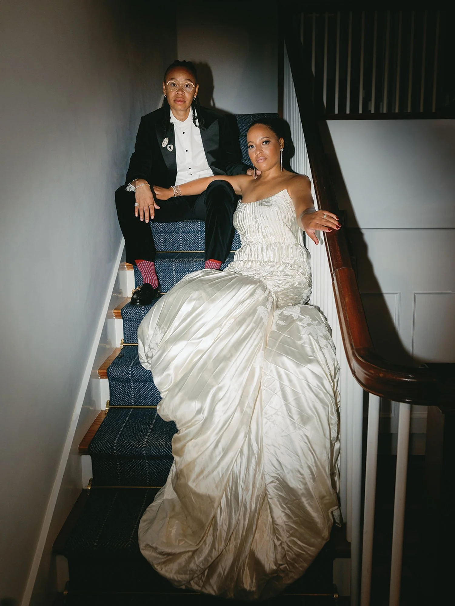 Two women on a staircase, one wearing a tuxedo and glasses, the other in an elegant white gown, both sitting and looking at the camera at Lambert's Cove Inn on Martha's Vineyard.
