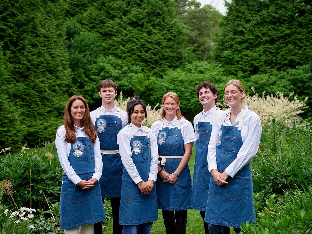Six people, five women and one man, standing outdoors in a garden with lush green trees in the background. They are all wearing white shirts and denim aprons, smiling at the camera at Restaurant Foster at Lambert's Cove Inn on Martha's Vineyard.