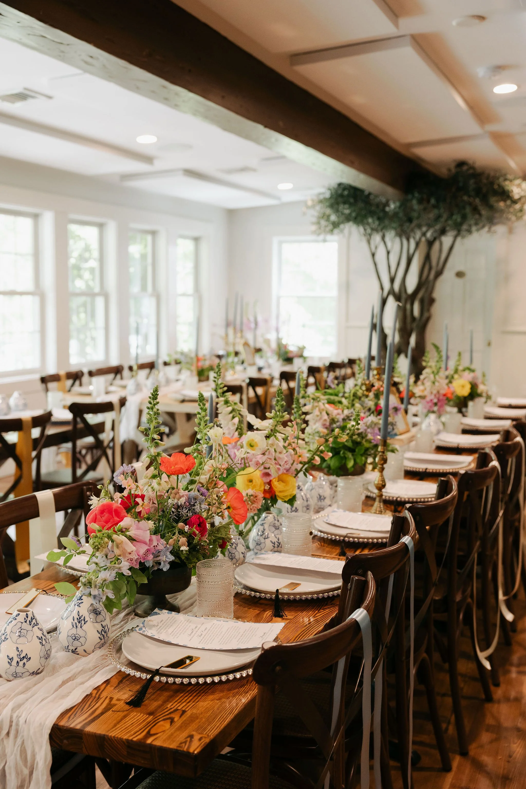 A decorated banquet table with floral centerpieces, tableware, and candles in a bright room with large windows and a tree in the background at Lambert's Cove Inn on Martha's Vineyard.