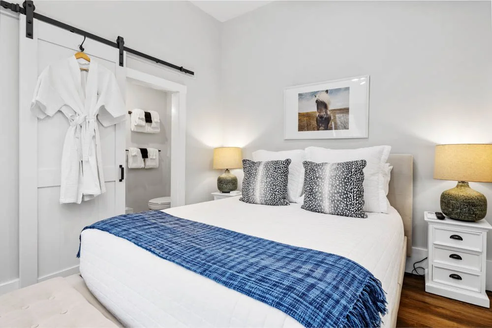 A neatly made bedroom with a white bed, blue blanket, and patterned pillows. Two nightstands with lamps flank the bed. A framed photo of a horse hangs above the bed. A white sliding barn door with a hanging white bathrobe and towels on hooks is visib