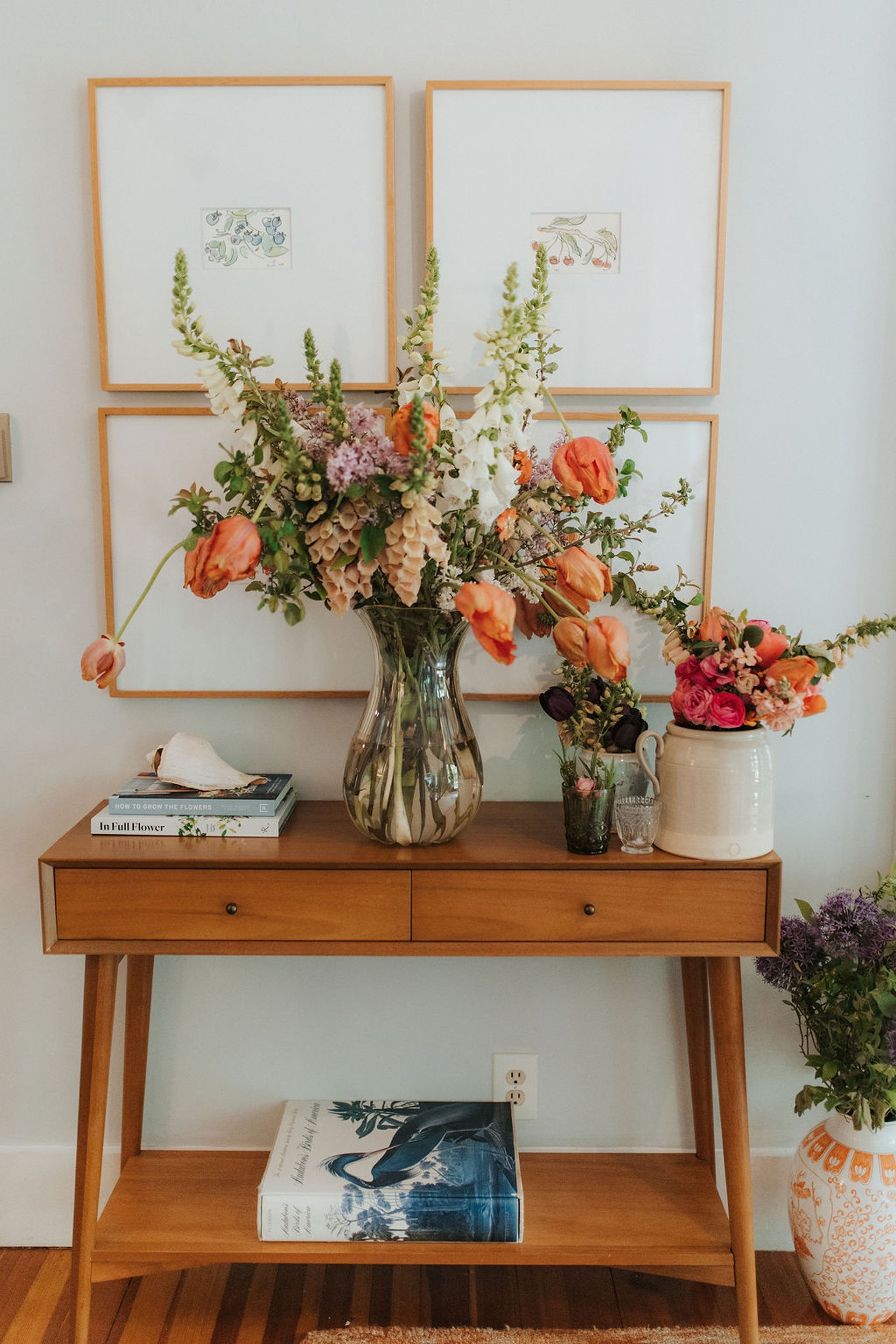 A wooden console table with two levels. The top level has a large vase with mixed flowers and smaller vases, and books. The lower level has a large book and a plant in a decorative ceramic vase. Framed artwork hangs on a white wall behind the table, with a small portion of a table or stool with a seashell on it visible.