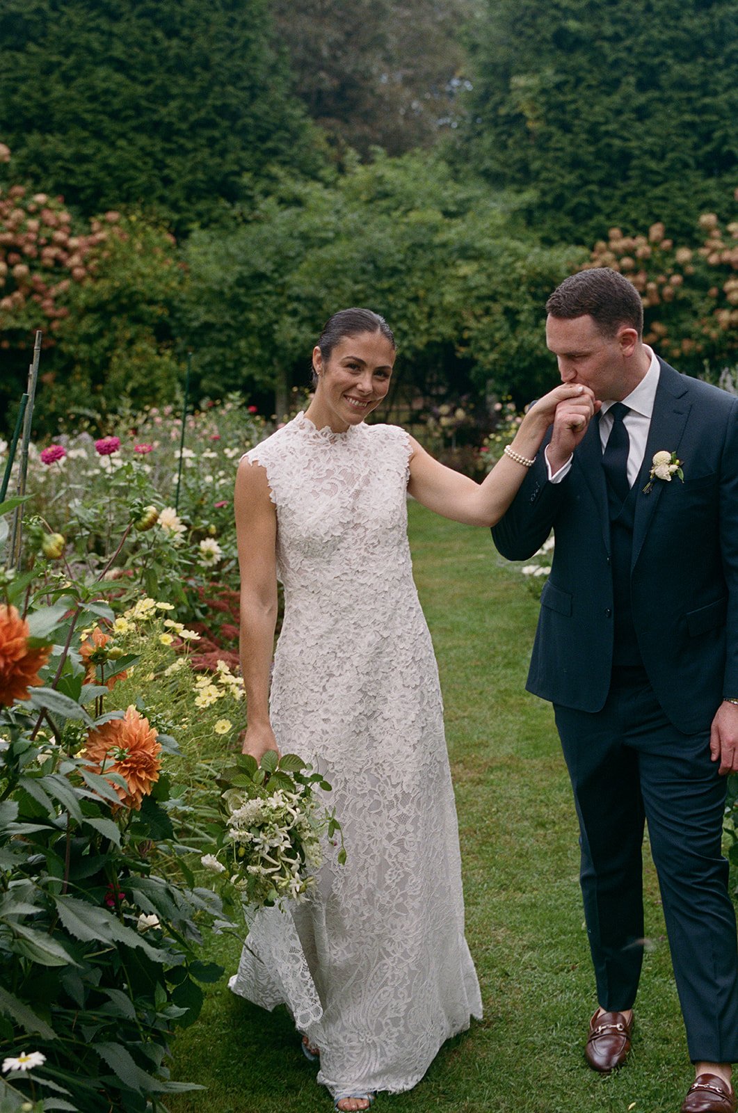 A woman in a white lace wedding dress is smiling and holding a bouquet, while a man in a dark suit kisses her hand in a garden with colorful flowers and greenery during a wedding at Lambert's Cove Inn on Martha's Vineyard.