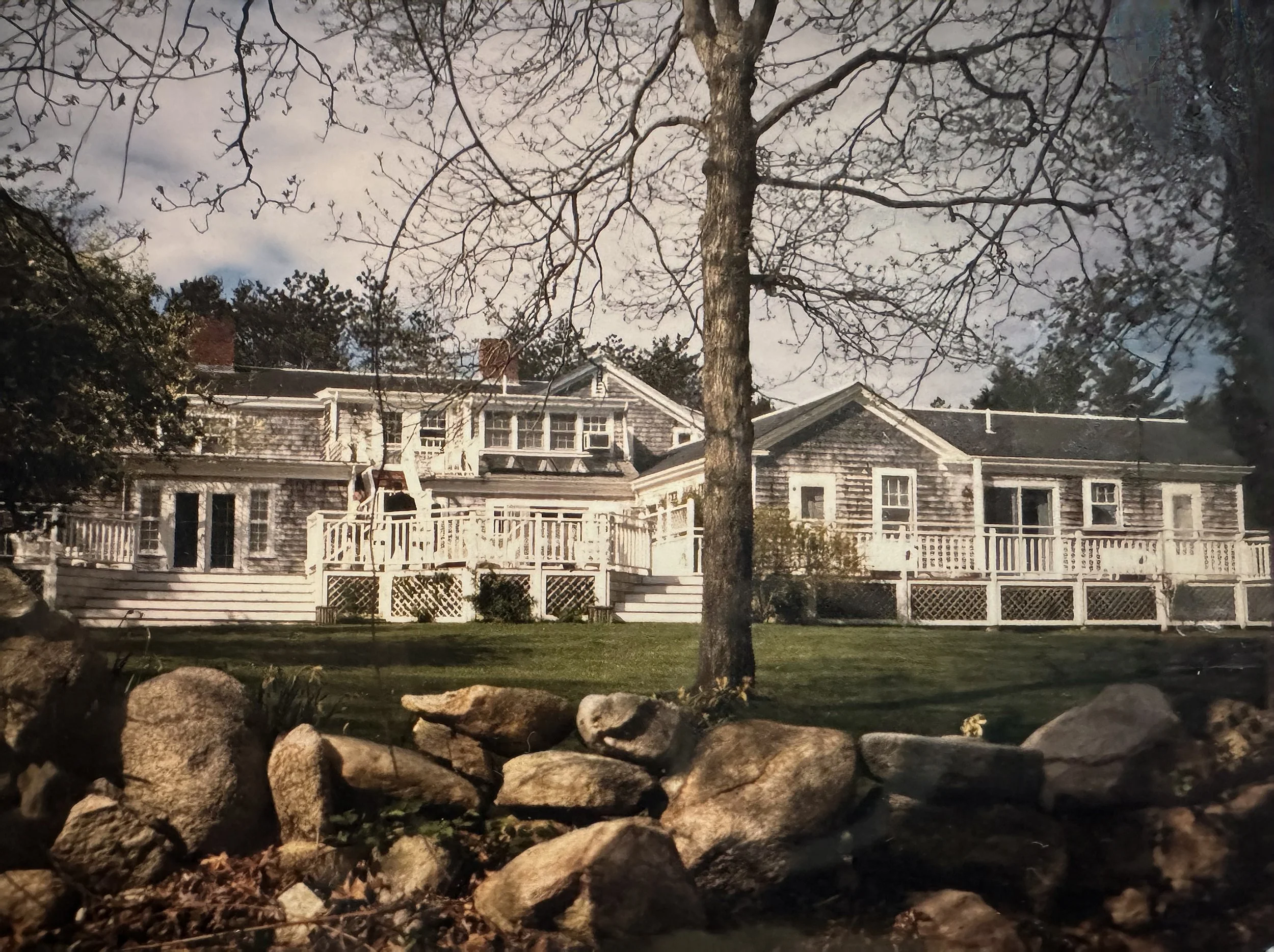 Nighttime view of a residential house with a deck, surrounded by a stone wall, with a large tree in the foreground and a cloudy sky above.