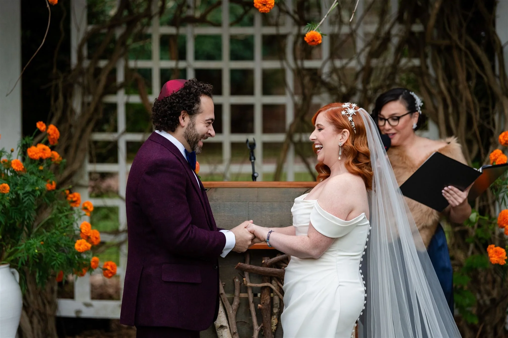 A couple exchanging wedding vows during a wedding ceremony outdoors at Lambert's Cove Inn on Martha's Vineyard, with a woman officiating and orange flowers hanging above.
