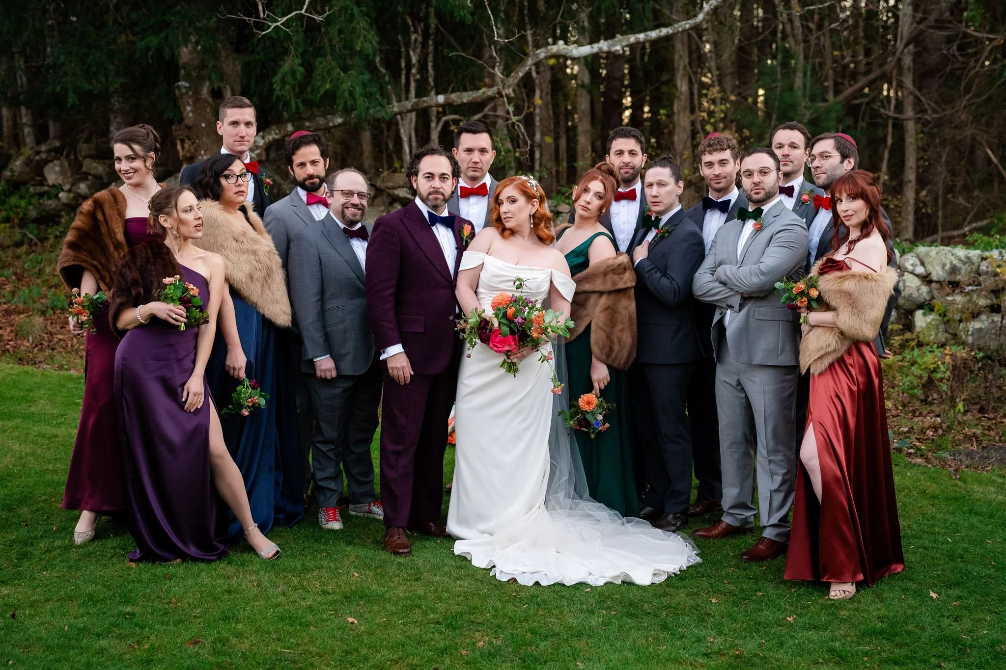 A large group of people dressed in formal attire, including tuxedos and evening gowns, gathered outdoors in front of a forested area for a wedding or special event. Some women are wearing fur stoles, and several people are holding small bouquets of f