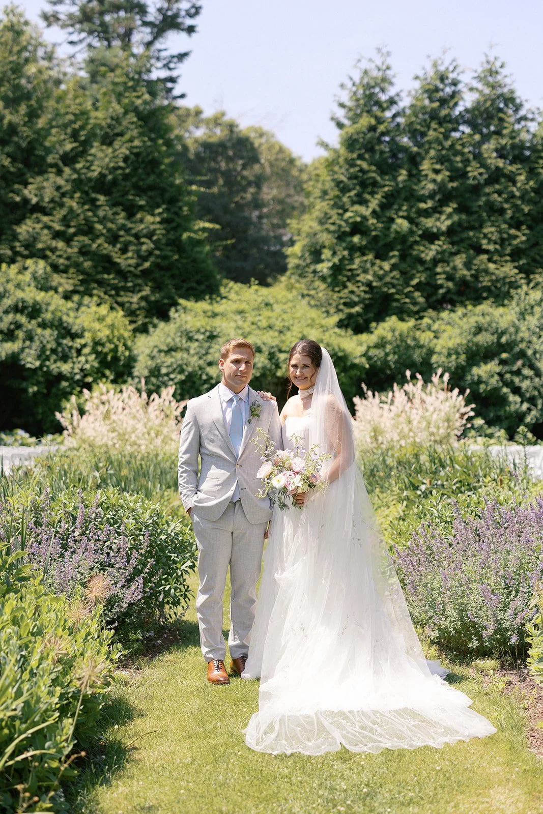 A bride and groom standing together outdoors in the English Garden at Lambert's Cove Inn on Martha's Vineyard on their wedding day, with lush green trees and colorful flowers in the background.