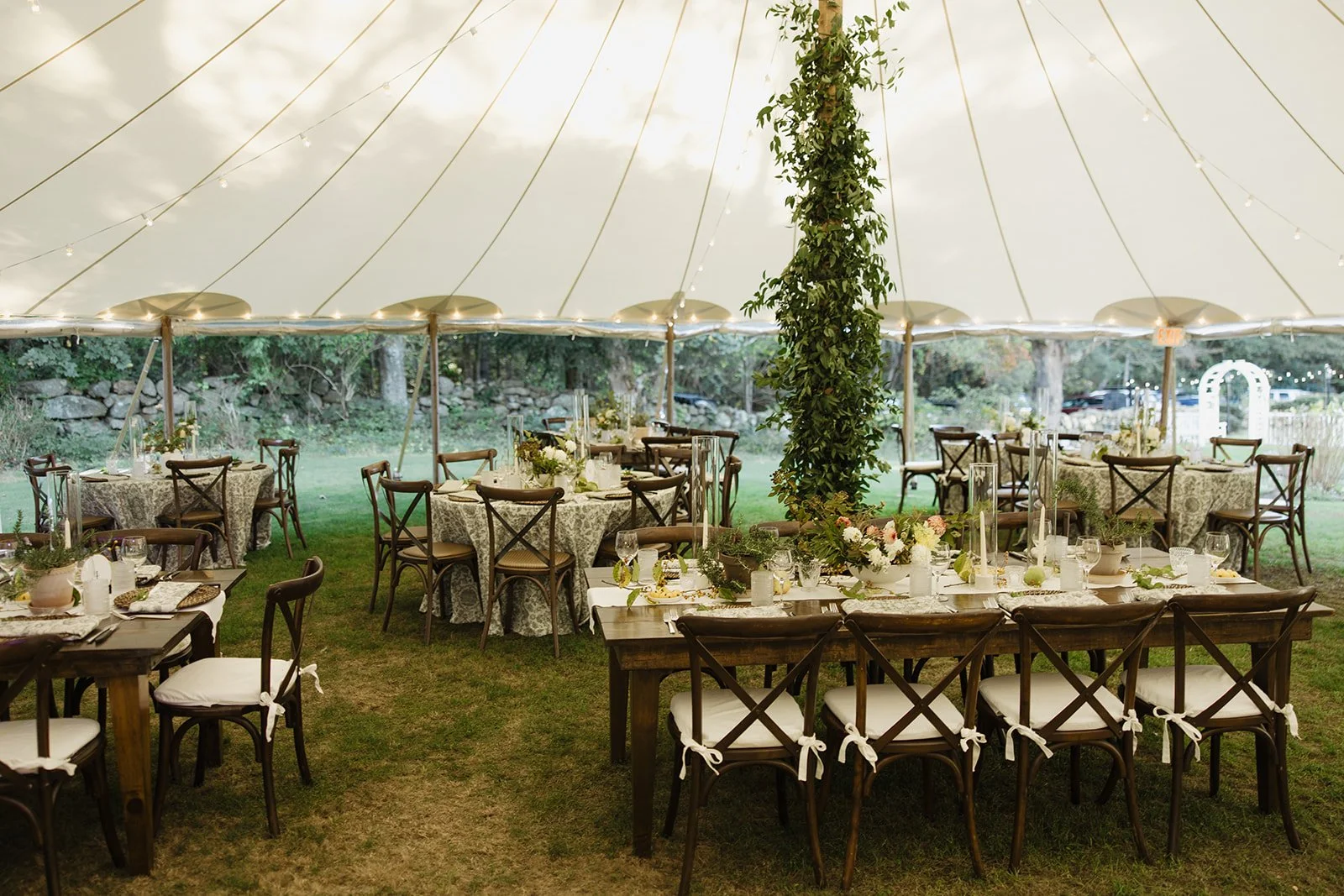 Elegant outdoor wedding reception at Lambert's Cove Inn on Martha's Vineyard under a large white tent with decorated tables, floral arrangements, and wooden chairs tied with white ribbons on grass.