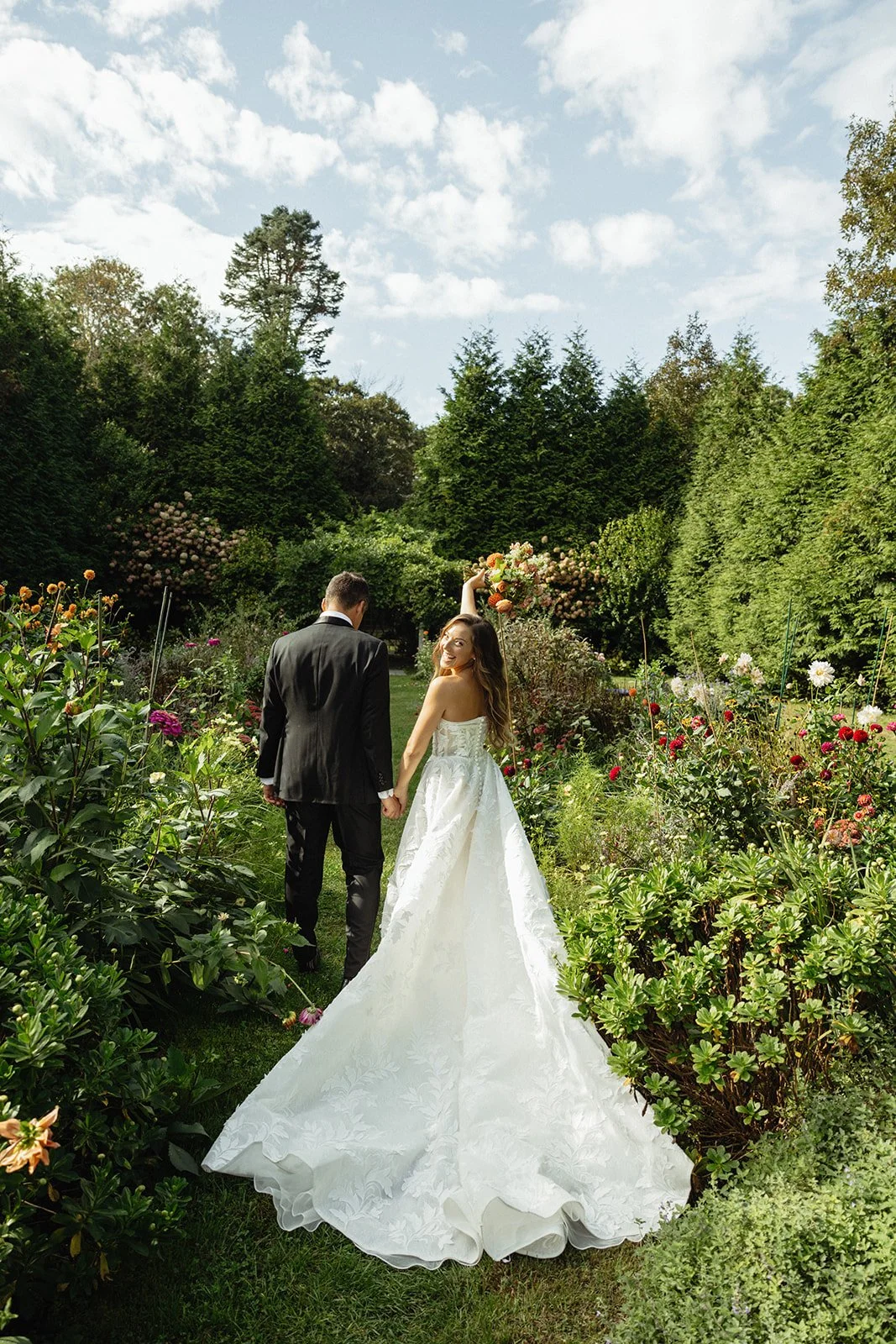 A bride and groom walking hand in hand through a lush garden with colorful flowers, greenery, and trees, under a partly cloudy sky during a wedding at Lambert's Cove Inn on Martha's Vineyard.