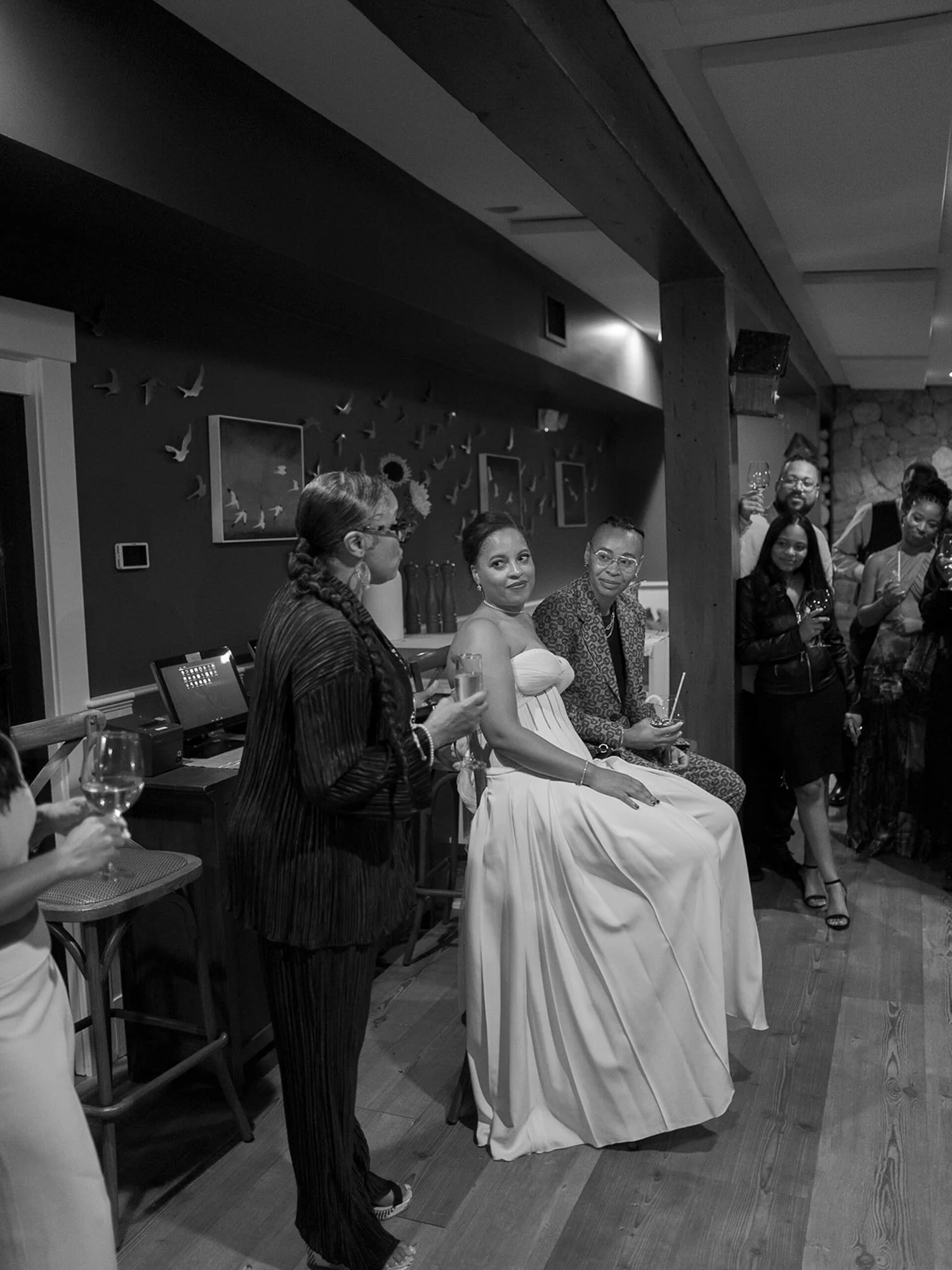 A bride in a strapless wedding gown sitting on a chair, surrounded by friends and family at a reception at Lambert's Cove Inn on Martha's Vineyard, with some holding glasses of wine, in a cozy indoor setting decorated with bird and flower wall art.