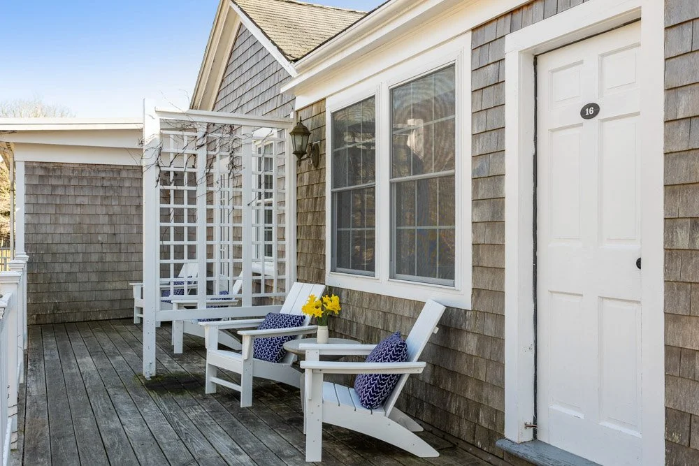 A porch with wooden flooring, gray shingled exterior walls, and white furniture, including two Adirondack chairs with pillows, a bench, and a plant with yellow flowers. There is a window with white trim and a door with a sign labeled '16'.