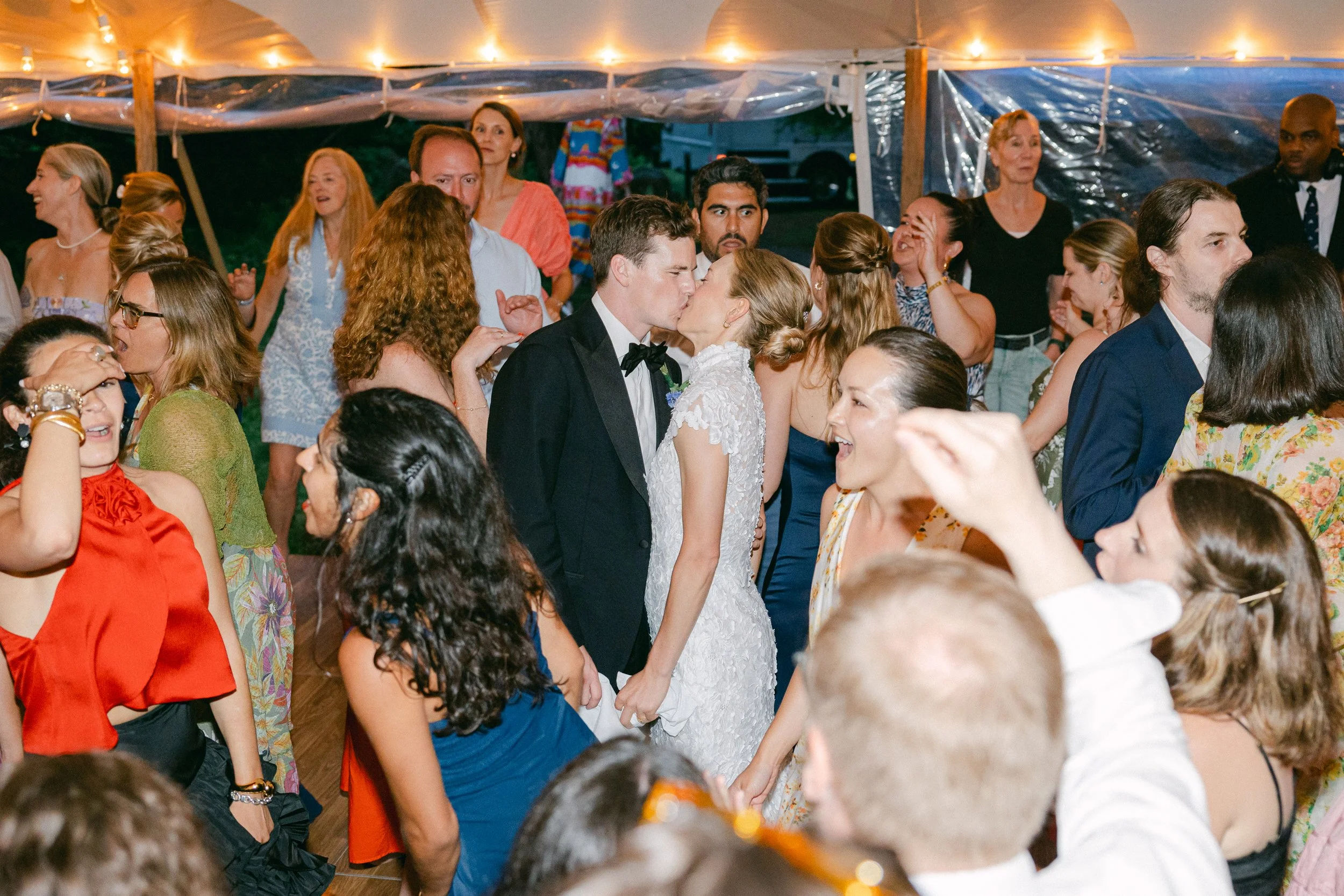 A bride and groom sharing a kiss while holding hands during a wedding at Lambert's Cove Inn on Martha's Vineyard surrounded by guests dancing and celebrating under a decorated tent.