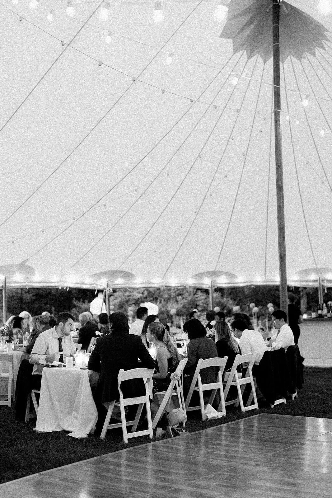 People sitting at a long table outdoors under a large umbrella during a wedding at Lambert's Cove Inn on Martha's Vineyard, with string lights overhead.