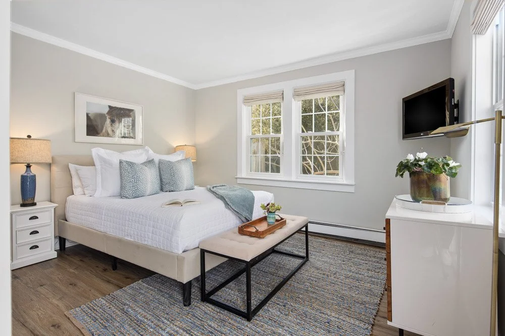 A bright bedroom with a large bed, white bedding, and decorative pillows. There are two bedside tables with lamps, a small bench at the foot of the bed, and a white dresser with a flowerpot on top. The room has light gray walls, two windows with blinds, a wall-mounted TV, and a framed picture of a dog.