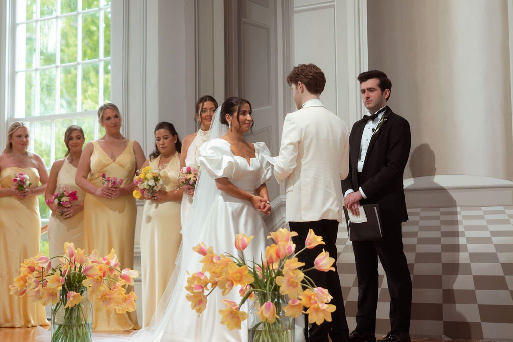 A wedding ceremony at Lambert's Cove Inn on Martha's Vineyard with a bride and groom exchanging vows, surrounded by bridesmaids and an officiant in a bright, elegant room with large windows and floral decorations.