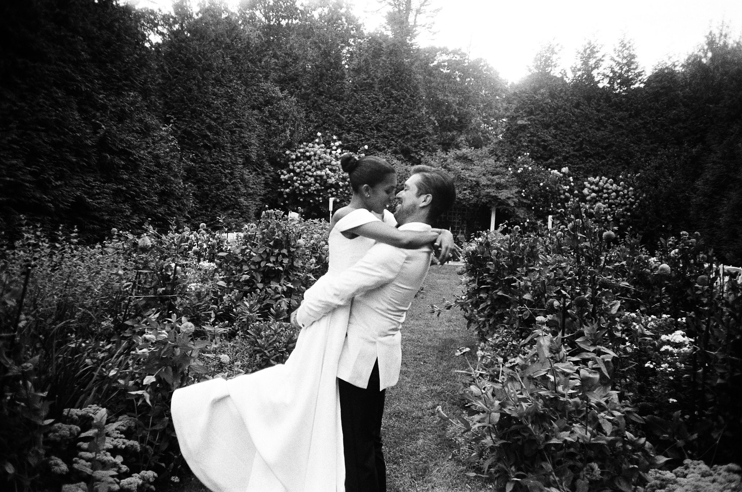 A couple dressed in formal wedding attire, with the bride in a white wedding gown and the groom in a suit, sharing a joyful moment in a garden with lush greenery and blooming flowers.