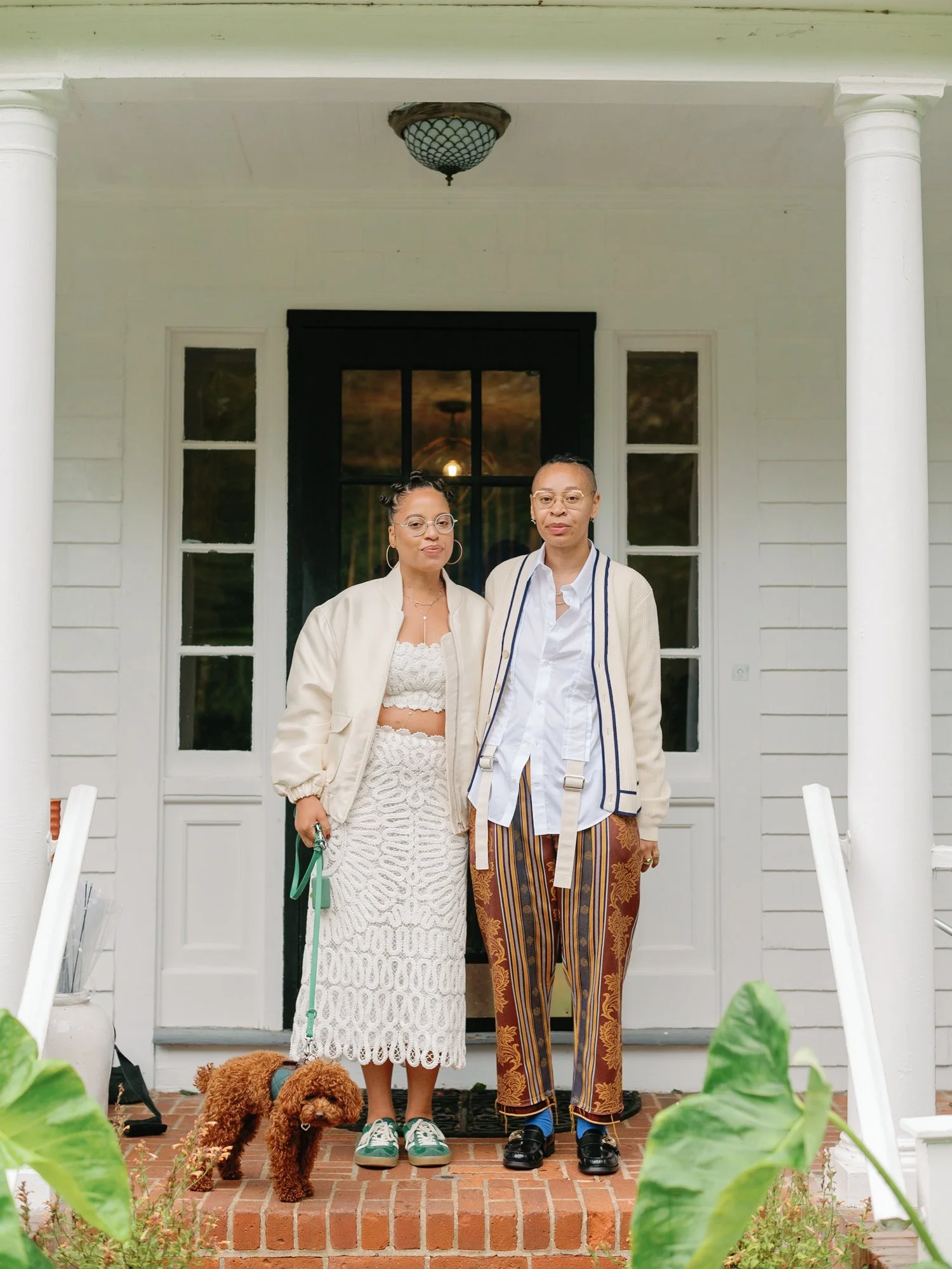 Two women standing on the front porch of a house, with a brown poodle dog sitting on the brick steps in front of them. The woman on the left is wearing glasses, hoop earrings, a beige jacket, a white lace dress, and green sneakers. The woman on the r
