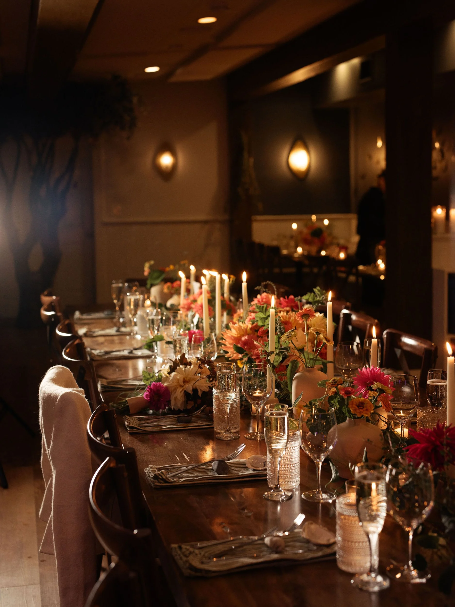 A table set for a formal dinner with floral centerpieces, candles, glassware, and place settings in a dimly lit room.