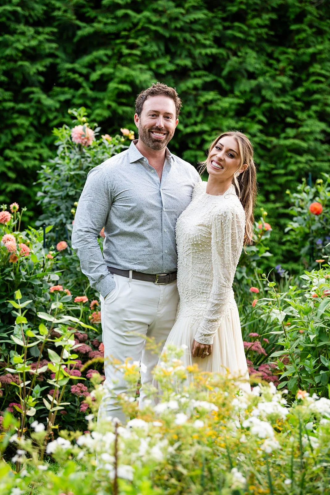 A smiling man and woman stand together outdoors in a garden  at Lambert's Cove Inn on Martha's Vineyard with colorful flowers and green foliage, appearing happy and affectionate.