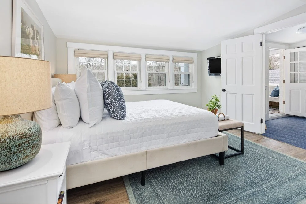 Bright bedroom with a large bed, white bedding, multiple pillows, a side table with a lamp, a wall-mounted TV, a potted plant, and a view into a sunroom or adjacent sitting area.