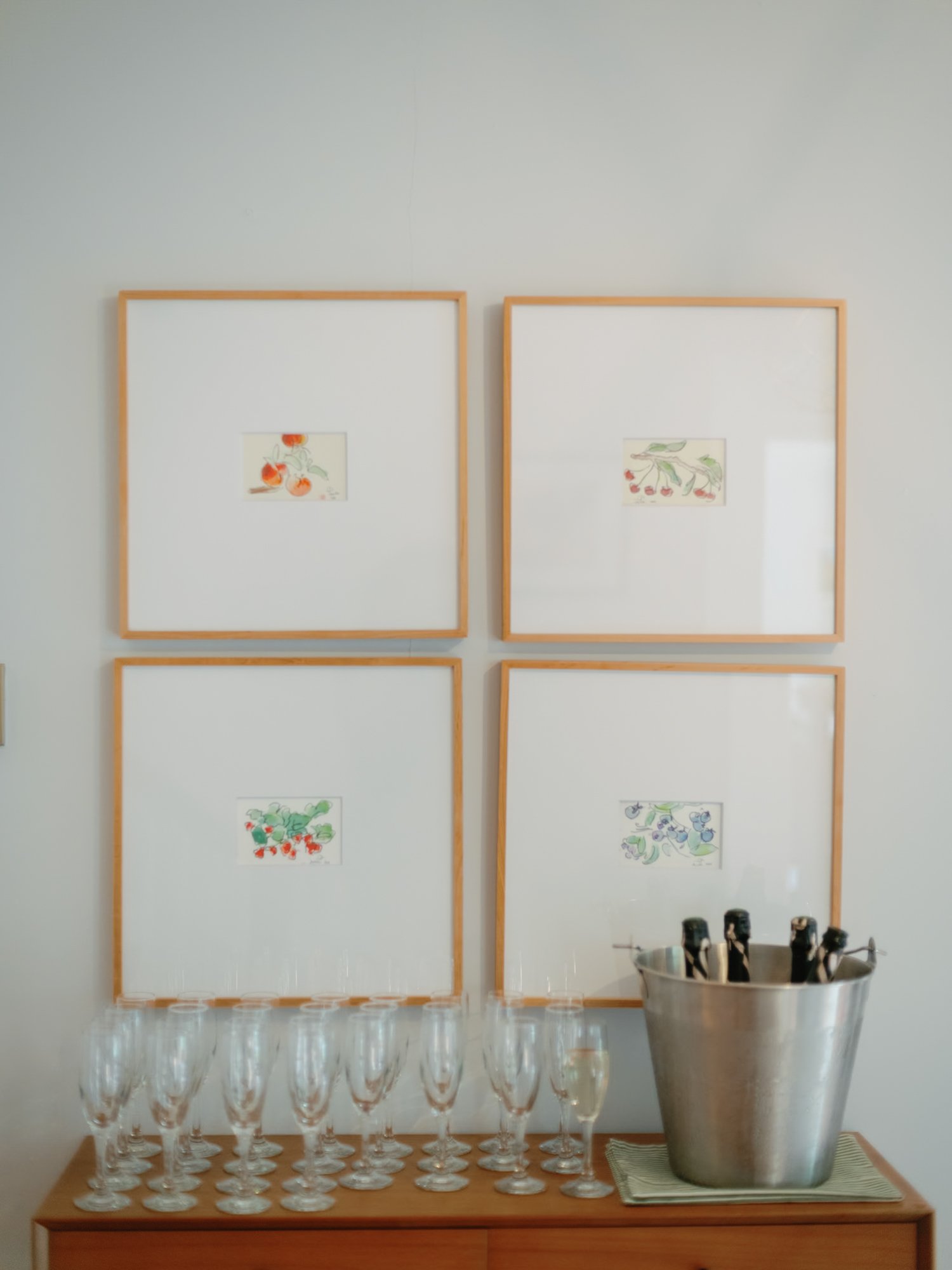 A wooden table holding fifteen empty champagne glasses arranged in rows, with a large metal bucket and chalkboard markers to the right. Behind the table are three large white-framed artworks with illustrations of tomatoes, cherries, and berries on a 